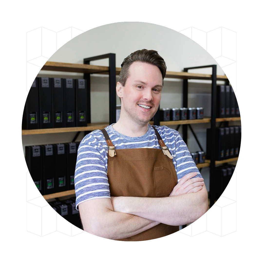 Photo of William Jollymore, the Chief Illumination Officer at Lunico Candle Co. He is standing with his arms crossed, with shelves of candles and reed diffusers in the background.